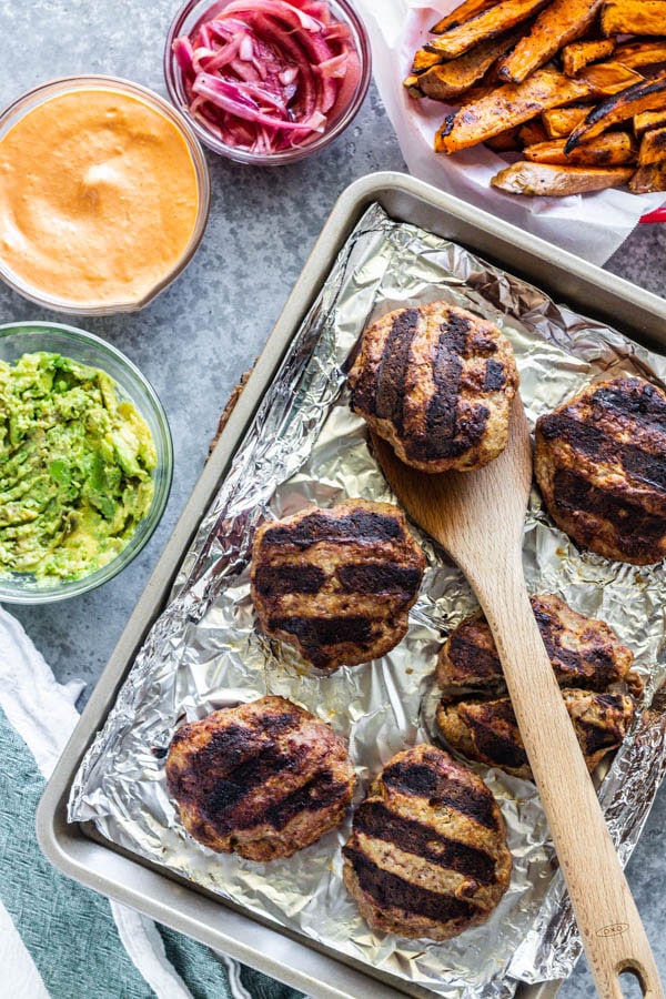 Burgers on a sheet pan with toppings around them.