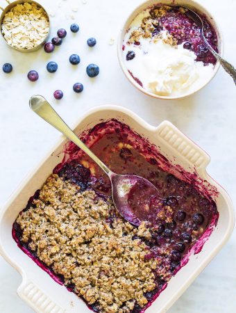 A pan of blueberry crisp with a bowl of ice cream , oats and blueberries.