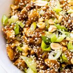 A bowl of sesame chicken with peanuts and scallions.