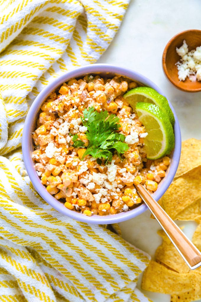 a bowl of elote with lime and chips around it.