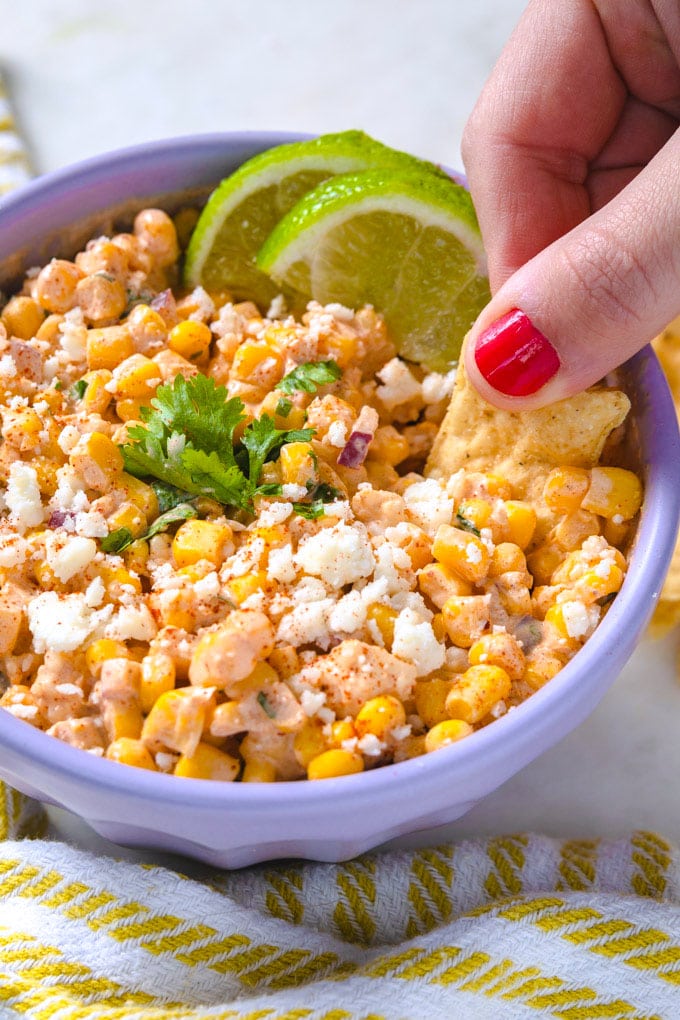 a hand dipping a tortilla chip into a bowl of Mexican street corn dip.