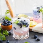 basil blueberry cocktail in a glass with ice cubes and blueberries around it.