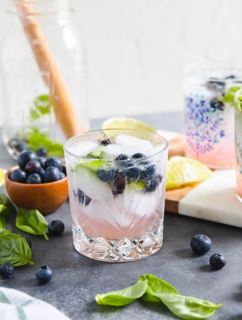 basil blueberry cocktail in a glass with ice cubes and blueberries around it.