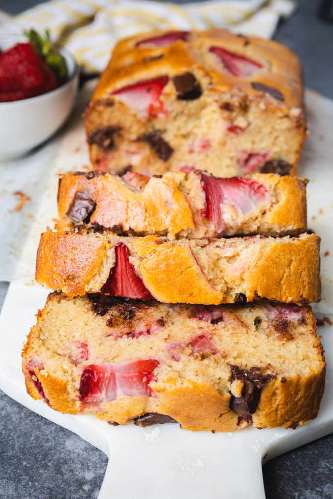 Sliced strawberry chocolate chip bread with strawberries next it on a cutting board. 