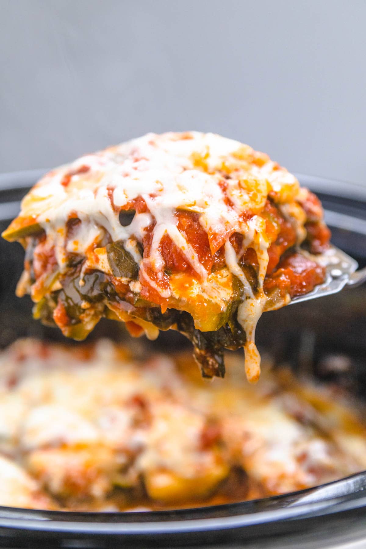 A piece of zucchini noodle lasagna dripping with cheese, on a spatula from a slow cooker. 