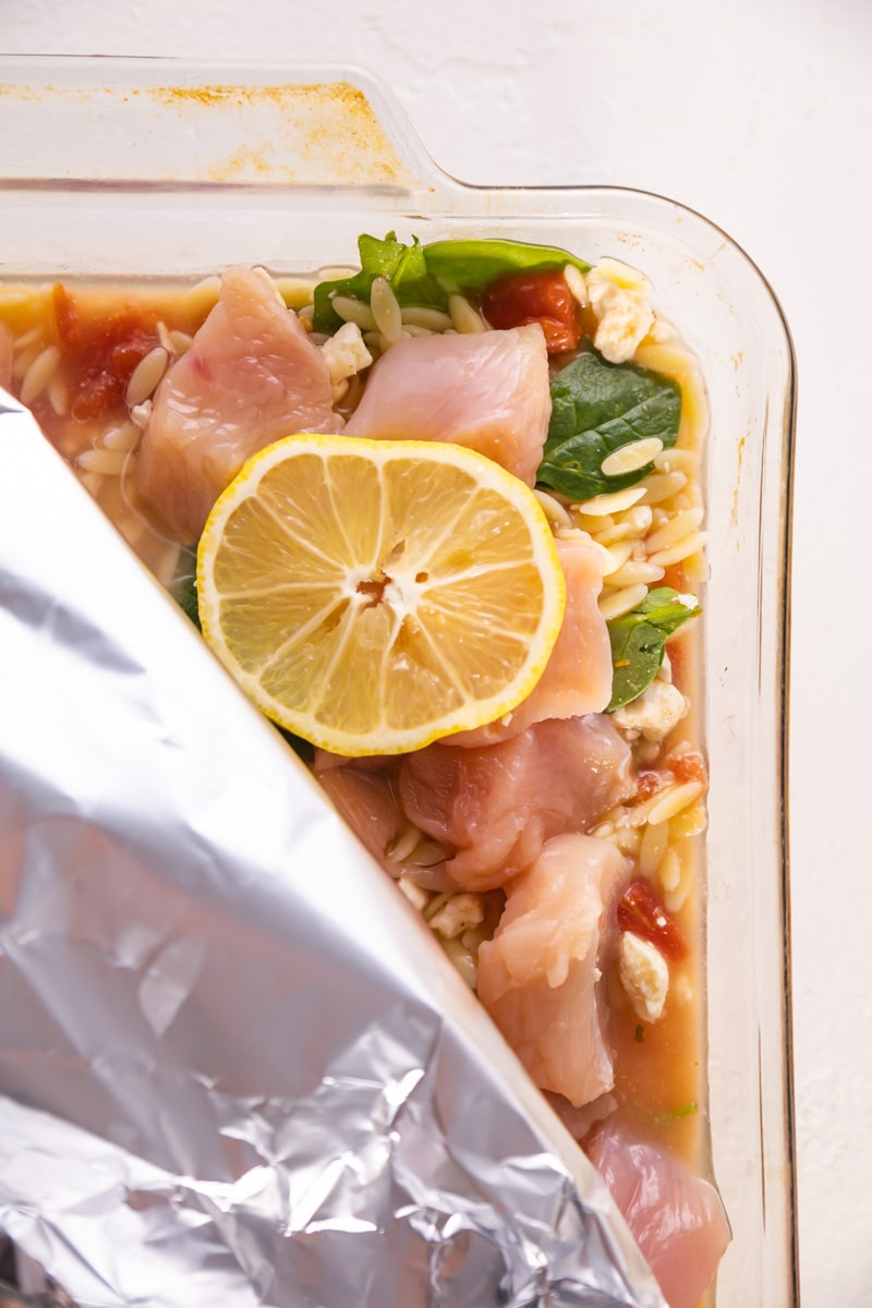 raw chicken on top of the casserole with foil and lemon on top ready to be baked in the oven.