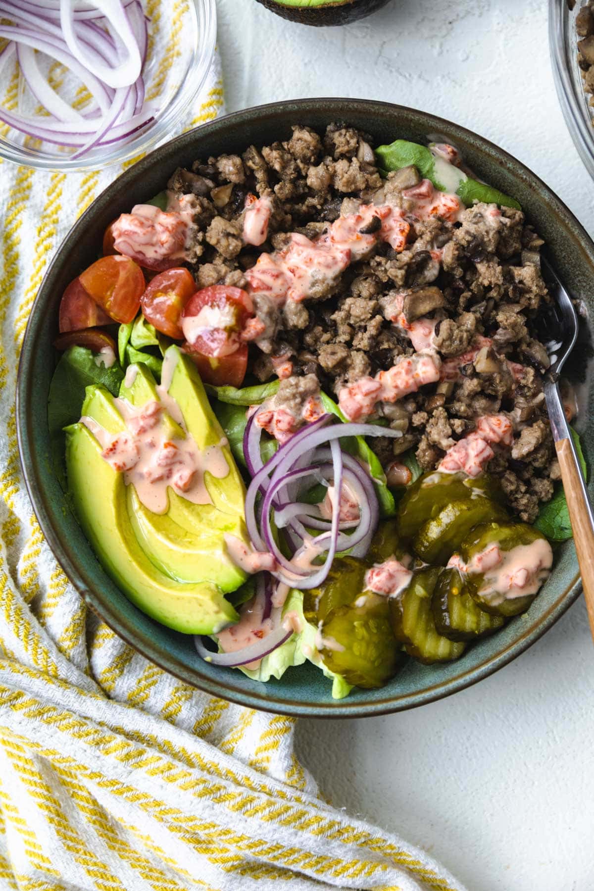 A BOWL FILLED WITH GROUND TURKEY, TOMATOES, PICKLES, AND OTHER BURGER BOWL TOPPINGS NEXT TO A NAPKIN WITH A FORK.