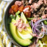 A CLOSE UP OF AVOCADO SLICES WITH AIOLI ON TOP INSIDE A BURGER BOWL WITH TOPPINGS.