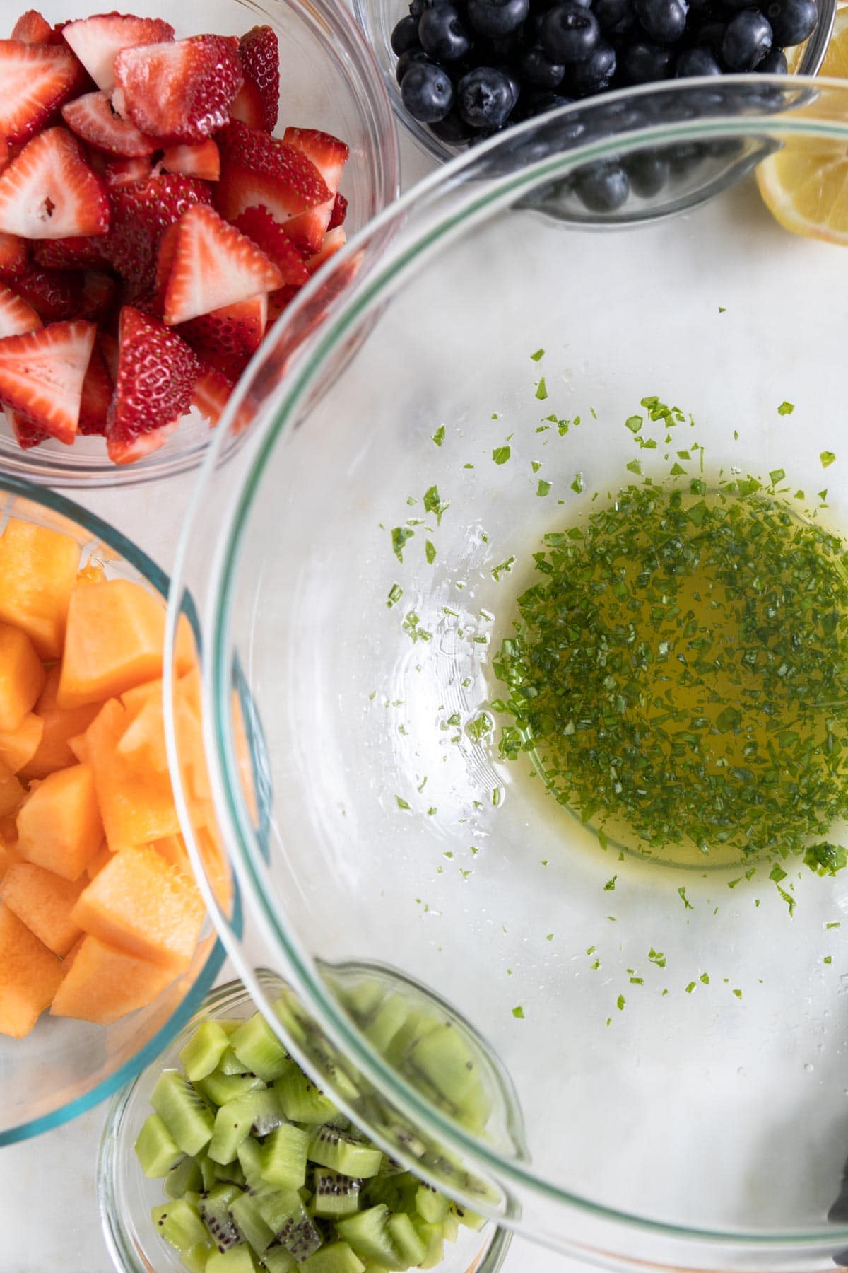 A bowl filled with minced basil, lemon juice and honey and bowls of fruit next to it.